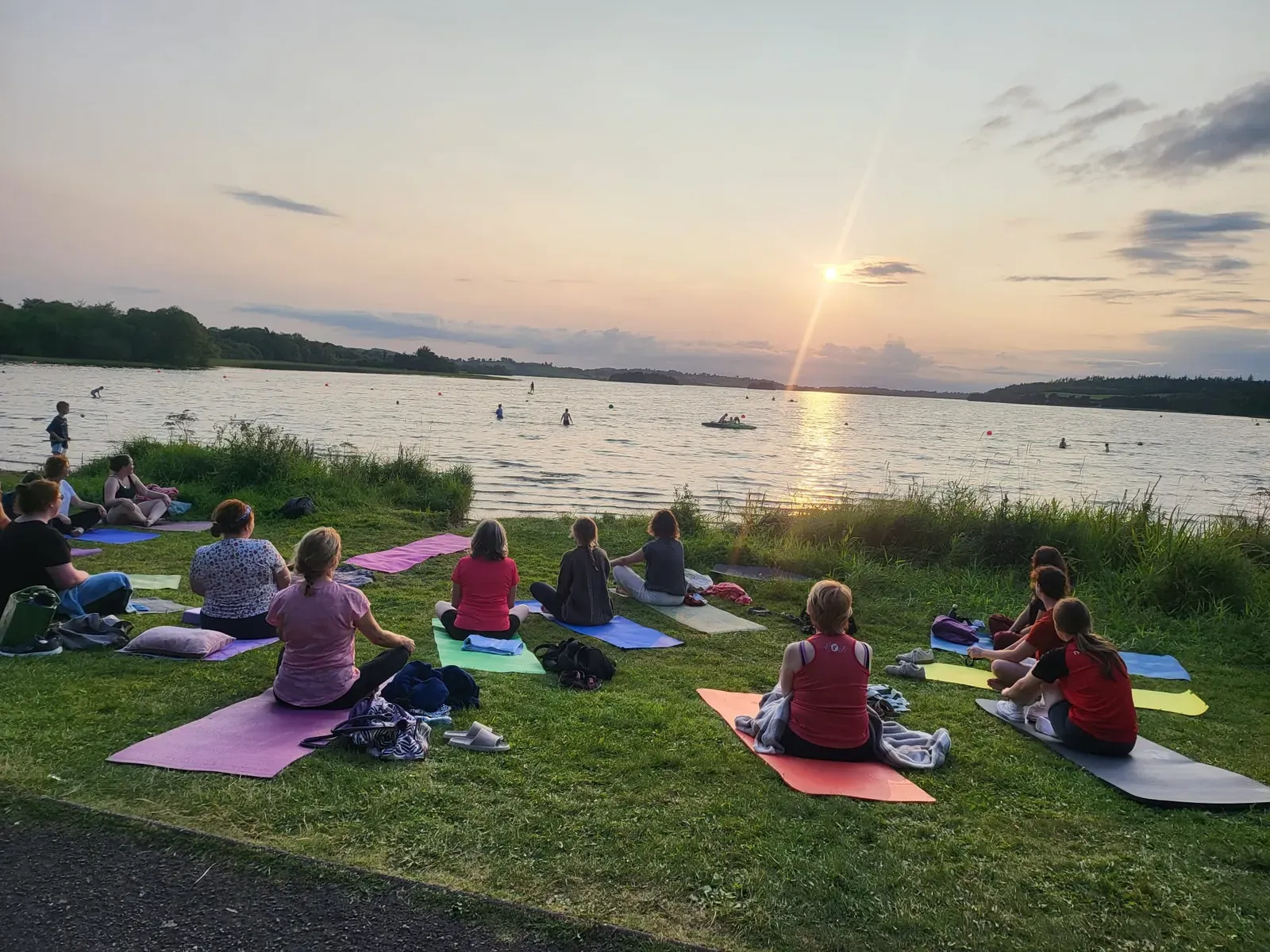 An outdoor yoga class sitting on mats by a lake at sunset on a summer evening in Meath